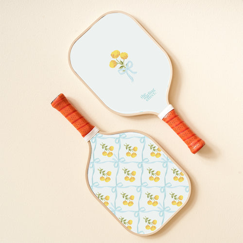 The-darling-effect's Marigold Lane Blue Pickleball Paddle features orange handles and lively designs: one with a yellow flower bouquet tied by a blue ribbon, the other with playful yellow flowers and blue ribbon patterns.