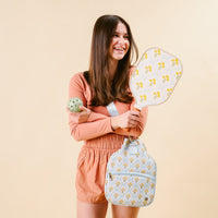 A smiling young woman in peach shorts and a pink top holds the-darling-effect’s Marigold Lane Blue Pickleball Paddle and a green ball, wearing a matching floral backpack, all set against a beige background.