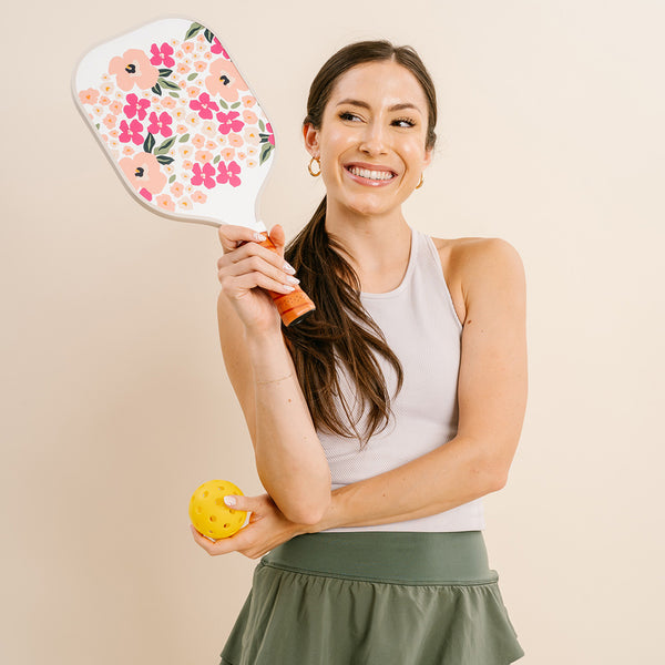 A woman in athletic wear smiles as she holds The Darling Effect’s Lively Flora Pink Pickleball Paddle and a yellow pickleball, proudly displaying her love for premium paddles.
