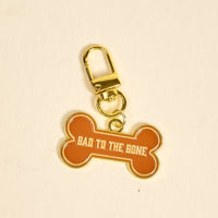 The Darling Effect’s Bad to the Bone Pawfect Collar Charm features an orange dog bone shape with gold edges, a gold clasp, and “BAD TO THE BONE” in bold white letters—a fun accessory for your pet’s collar or keychain.