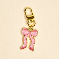 The Darling Effect Bow Pink Pawfect Collar Charm is a gold keychain featuring a pink, gold-outlined bow with white dots—perfect as an adorable pet accessory or collar charm. Pictured on a light beige background.