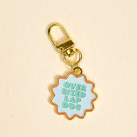 The Darling Effect’s Oversized Lap Dog Pawfect Collar Charm is a gold keychain with a wavy light blue tag reading OVER SIZED LAP DOG—perfect for adding playful pet style. Shown on a plain beige background.