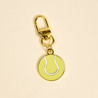 The Darling Effect's Tennis Ball Pawfect Collar Charm features a gold keychain with a yellow and white tennis ball—ideal for adding playful style to your keys or as a trendy accessory for your pet’s collar.