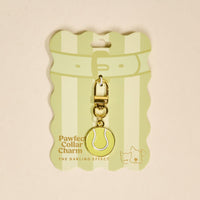 The Darling Effect's Tennis Ball Pawfect Collar Charm is a gold, tennis ball-shaped charm with clasp, displayed on striped green packaging featuring cute cat and dog illustrations—perfect for playful pet style lovers.