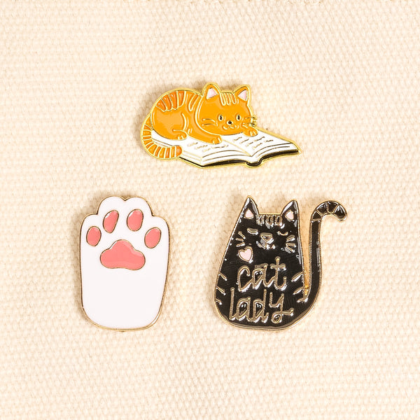 Cool Cat The Pin Pack by The Darling Effect features three expressive pins: a cat on a book, a pink and white cat paw, and a black cat with "cat lady" script—a charming addition to any pin collection.