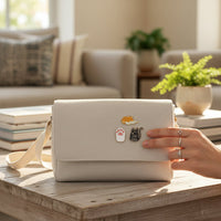 A hand with silver rings touches a cream-colored purse adorned with The Darling Effect’s Cool Cat The Pin Pack on a wooden coffee table, surrounded by plants and books in a sunlit living room.