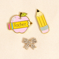 Classroom Charm The Pin Pack by The Darling Effect features three enamel pins: a pink apple with a yellow "Teacher" banner, a yellow pencil with pink eraser, and a rhinestone-studded bow—perfect for any collection.
