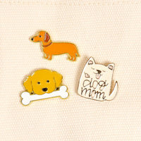 Dog Days The Pin Pack by The Darling Effect features three playful pins on cream fabric: an orange dachshund, a yellow dog with a bone, and a white cat with "dog mom" text—perfect for animal lovers!.