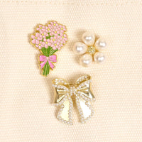 The Darling Effect's Floral Fancy The Pin Pack includes three collectible pins—a pink floral bouquet, a pearl flower, and a pearl-accented gold bow—to add elegance to any outfit or bag.