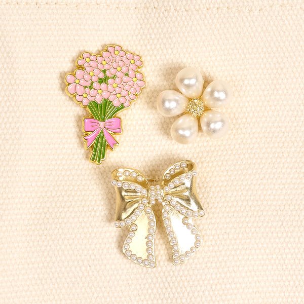The Darling Effect's Floral Fancy The Pin Pack includes three collectible pins—a pink floral bouquet, a pearl flower, and a pearl-accented gold bow—to add elegance to any outfit or bag.