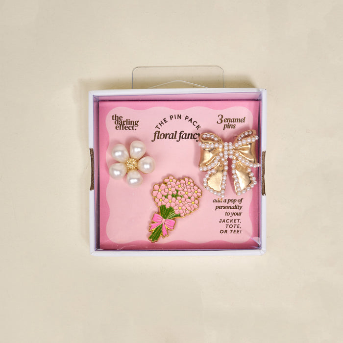 The Darling Effect's Floral Fancy The Pin Pack features three collectible pins—a pearl flower, gold bow with pearl details, and pink bouquet—to add playful charm to your jacket, tote, or tee.