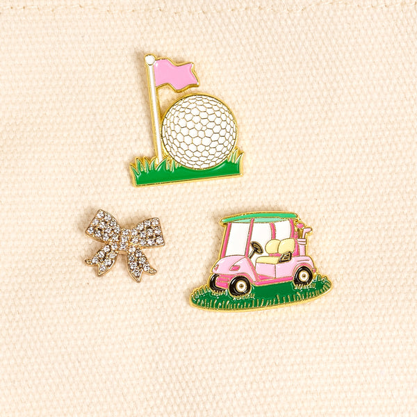 Golf Par-Tee The Pin Pack by The Darling Effect features three pins on cream fabric: a shiny silver bow, a pink and white golf cart on grass, and a golf ball near a pink flag—perfect for any curated pin collection.