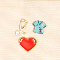 Nurse Life The Pin Pack by The Darling Effect features three pins on a cream background: a white and gold stethoscope, a teal scrubs top with red cross, and a bold red heart with gold border—perfect for any pin collection.