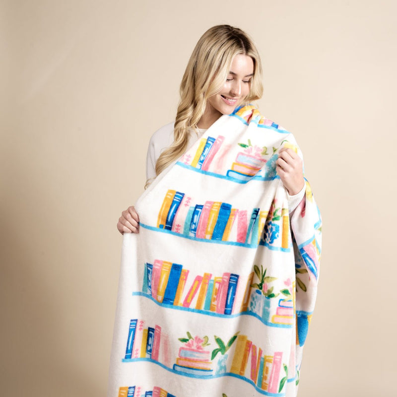 A woman with long blonde hair smiles while holding The Darling Effect's Book Shelfie Snuggle Up Plush Blanket, which features illustrated books and plants on shelves, against a neutral background.