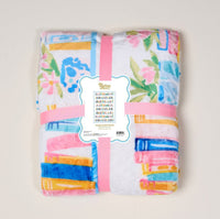 The Darling Effect Book Shelfie Snuggle Up Plush Blanket is a folded, cozy throw with colorful floral and geometric patterns, wrapped in a pink ribbon and decorative label, displayed on a light background.