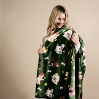 A smiling woman with long blonde hair wraps herself in The Darling Effect’s Dog Days Snuggle Up Plush Blanket, featuring playful cartoon dogs, bones, and tennis balls, against a plain beige background.