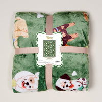The Darling Effect’s Dog Days Snuggle Up Plush Blanket is green with playful cartoon dogs, rolled and tied with a beige ribbon. A front label shows the blanket pattern and product details.