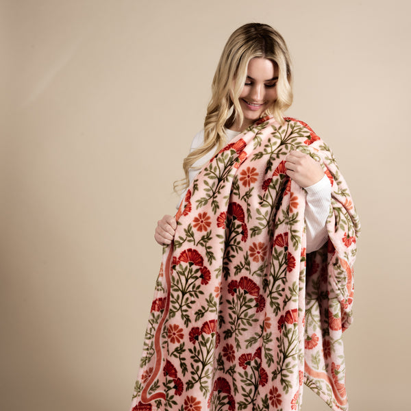 A person with long blonde hair in a white top smiles while holding and admiring The Darling Effect's Everbloom Dusty Rose Snuggle Up Plush Blanket, featuring red and pink flowers with green leaves, against a neutral background.