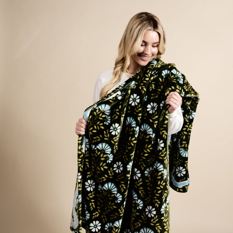 A woman with long blonde hair smiles while holding The Darling Effect's Everbloom Forest Green Snuggle Up Plush Blanket, featuring blue and white floral designs, against a beige background.