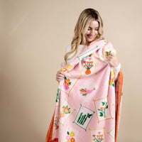 A young woman with blonde hair smiles as she holds up The Darling Effect's It's Mahj Time Snuggle Up Plush Blanket, a soft pink fleece adorned with framed squares featuring vibrant floral and bird illustrations.