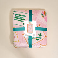The Darling Effect’s "It’s Mahj Time Snuggle Up Plush Blanket," featuring colorful geometric and abstract patterns in pink, is folded with a teal ribbon and branded label, offering cozy comfort against a plain light background.
