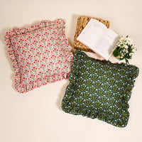 The Darling Effect Everbloom Forest Green Scalloped Square Throw Pillow rests on a light surface beside another pillow, a woven mat, an open book, and a small bouquet of white flowers for a cozy decorative touch.