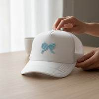 A person adjusts a white mesh baseball cap featuring The Darling Effect's Bow Blue Mix & Match Iron-On Patch, ready for further customization, on a light wooden table beside a white cup.