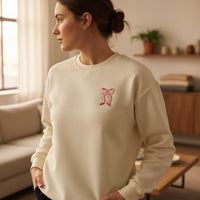 A woman indoors wears a cream sweatshirt featuring The Darling Effect's Bow Pink Mix & Match Iron-On Patch. She looks to the side in a softly lit room with a sofa and plant-filled shelves in the background.