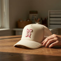 A hand holds the brim of a beige baseball cap topped with The Darling Effect's Bow Pink Mix & Match Iron-On Patch, resting on a wooden table in a softly lit room—perfect inspiration for customizing jackets or other items.