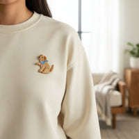 Close-up of The Darling Effect’s Dog Mix & Match Iron-On Patch, featuring an embroidered golden retriever puppy in a blue bandana. Customize your cream sweatshirt or other items with this patch. Cozy living room background softly blurred.