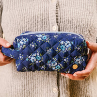 A person in a light gray sweater holds the-darling-effect's Endless Daydream Navy Essentials Quilted Cosmetic Pouch—a navy blue makeup bag with a blue floral pattern, wipeable interior, and a small round gold emblem.