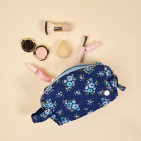 The-darling-effect’s Endless Daydream Navy Essentials Quilted Cosmetic Pouch, with a wipeable interior and zipper, rests among makeup must-haves—a brush, compact powder, sponge, lipstick, and gloss—on a beige background.