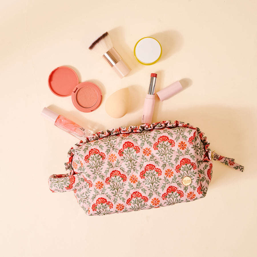 The Darling Effect’s Everbloom Dusty Rose Essentials Quilted Cosmetic Pouch, featuring a floral pattern and zipper, sits open on a light beige surface with makeup items nearby. Its wipeable interior ensures easy cleaning.