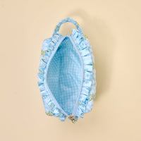 The Darling Effect Oh So Fancy Blue Essentials Quilted Cosmetic Pouch features light blue floral exterior, ruffled details, a wipeable gingham interior lining, and a top handle, shown open from above on a beige background.