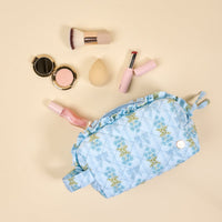 The Darling Effect's Oh So Fancy Blue Essentials Quilted Cosmetic Pouch, featuring ruffles and floral quilting, is displayed on a beige background surrounded by makeup items like a brush, sponge, lipstick, nail polish, blush, and compact.