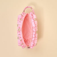 The Darling Effect’s Oh So Fancy Pink Essentials Quilted Cosmetic Pouch features pink ruffled quilting, floral embroidery, a zipper closure, and a pink and white gingham-lined interior, shown partially open on a beige background.