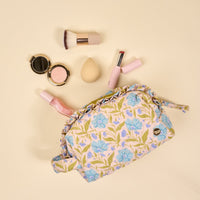 The Darling Effect's Poppin' Petals Aqua Essentials Quilted Cosmetic Pouch, featuring blue floral patterns, is open on a beige surface surrounded by makeup essentials like a brush, compact powder, sponge, lipstick, and nail polish.