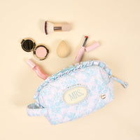 The Darling Effect’s Mrs. Something Blue Essentials Quilted Cosmetic Pouch, in light blue floral with a "MRS" label, is displayed on a beige background surrounded by a makeup brush, compact powder, beauty sponge, lipstick, and lip gloss.