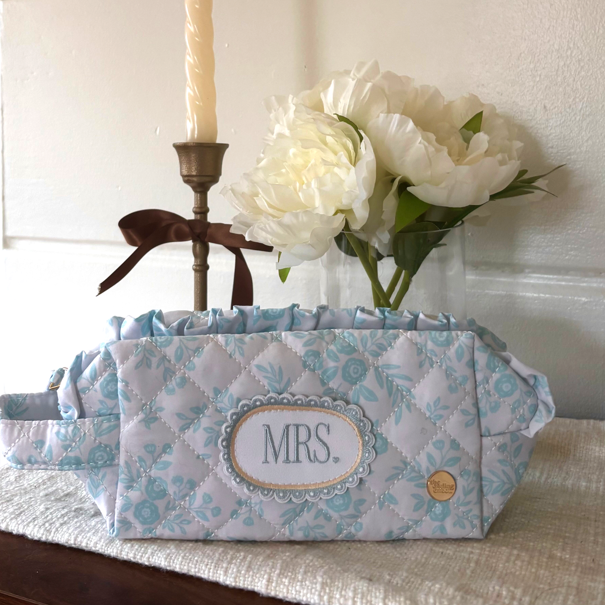 The Darling Effect’s Mrs. Something Blue Essentials Quilted Cosmetic Pouch, featuring a floral pattern and MRS. embroidery, sits on a mat with a candle and white peonies in the background—ideal for carrying your beauty essentials.