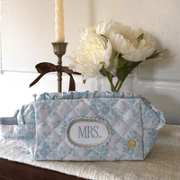 The Darling Effect’s Mrs. Something Blue Essentials Quilted Cosmetic Pouch, featuring a floral pattern and MRS. embroidery, sits on a mat with a candle and white peonies in the background—ideal for carrying your beauty essentials.