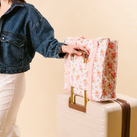 A person in a denim jacket holds the-darling-effect’s Endless Daydream Cream The Everyday Quilted Tote Bag with pink trim, perfect for daily use. The floral tote rests on a cream suitcase.