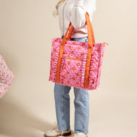 A woman in a white sweater and blue jeans carries The Darling Effect's Poppin' Petals Hot Pink The Everyday Quilted Tote Bag, featuring orange handles, ruffle details, an exterior zip pocket, and styled casually with sneakers.