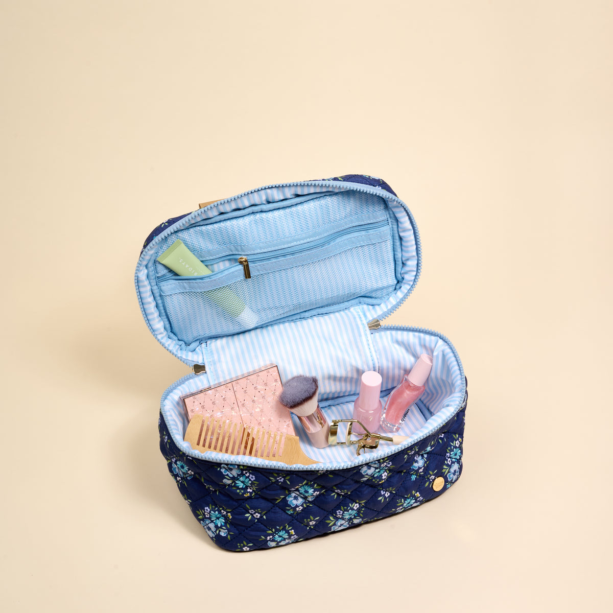 The Endless Daydream Navy Glam & Go Quilted Cosmetic Bag from the-darling-effect features a blue floral design with a light blue striped lining. Shown unzipped on a beige background, it holds essentials like brushes, a palette, lip gloss, and a green tube—making it the perfect travel makeup bag.