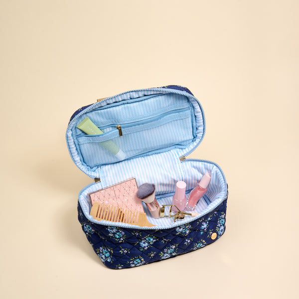 The Endless Daydream Navy Glam & Go Quilted Cosmetic Bag from the-darling-effect features a blue floral design with a light blue striped lining. Shown unzipped on a beige background, it holds essentials like brushes, a palette, lip gloss, and a green tube—making it the perfect travel makeup bag.