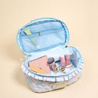 The Darling Effect's Bride Something Blue Glam & Go Quilted Cosmetic Bag is open, showing nail polishes, a powder brush, pink pouch, cream tube, and hair clip inside—all set against a beige background.