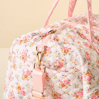 A close-up of the-darling-effect Endless Daydream Cream On-the-Go Quilted Weekender Bag with a pink and orange floral pattern, gold hardware, pink handles, and an adjustable strap, set against a plain beige background.