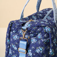 A close-up of the-darling-effect Endless Daydream Navy On-the-Go Quilted Weekender Bag, featuring a floral pattern, light blue zipper and strap with gold hardware, resting on a light surface against a beige background.