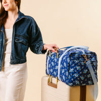A woman in a denim jacket and white pants stands next to a suitcase, resting her hand on the-darling-effect’s Endless Daydream Navy On-the-Go Quilted Weekender Bag, which sits atop the suitcase against a beige background.