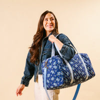 A woman with long brown hair in a denim jacket and light pants smiles while carrying the-darling-effect's Endless Daydream Navy On-the-Go Quilted Weekender Bag, featuring a floral pattern, over her shoulder against a beige background.