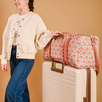 A person in a cream jacket and blue jeans stands by a large cream suitcase with The Darling Effect's Everbloom Dusty Rose On-the-Go Quilted Weekender Bag, featuring an adjustable shoulder strap, resting on top against a beige background.